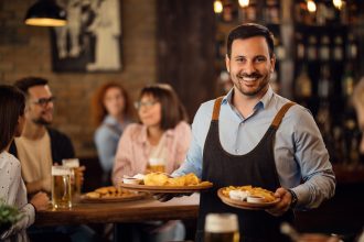 Happy waiter holding plates with food and looking at camera while serving guests in a restaurant.